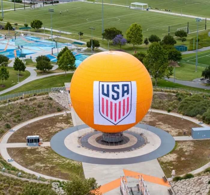 Large orange sphere with the USA soccer logo, set in a park with sports fields.