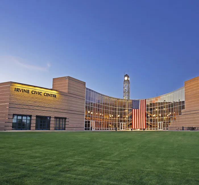Modern Irvine Civic Center with glass facade, clock tower, and American flag over green lawn.