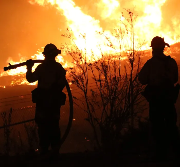 Silhouetted firefighters stand before a massive wildfire at night.