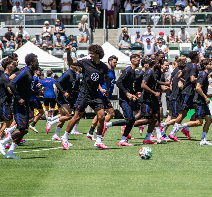 Soccer players in dark jerseys and shoes run across a green field during practice.