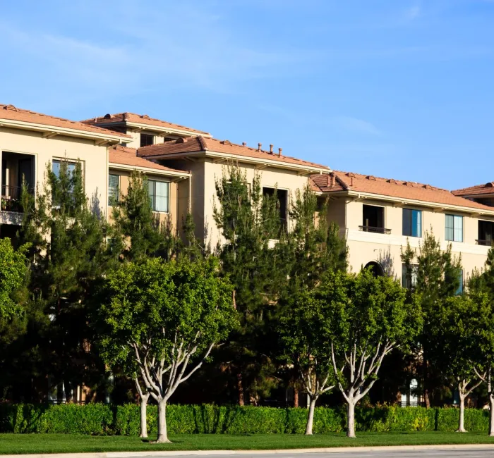 Modern tan apartment building with red tile roofs and lush green trees.