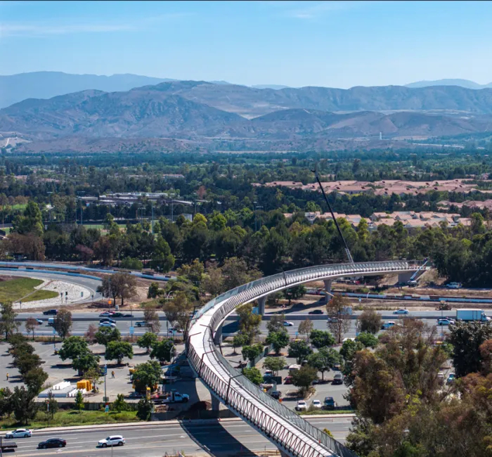 Elevated train track over a highway and parking lot, with green fields and mountains behind.