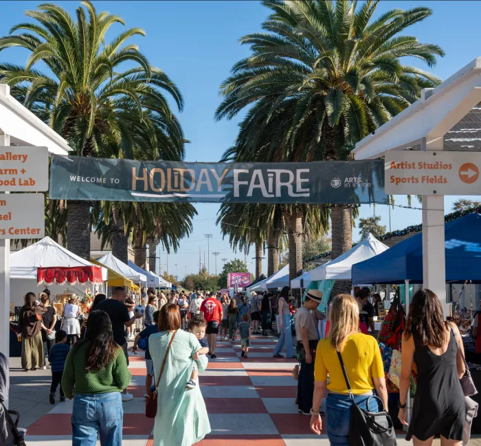 Photograph: A bustling outdoor Holiday Faire with people, market stalls, and palm trees.