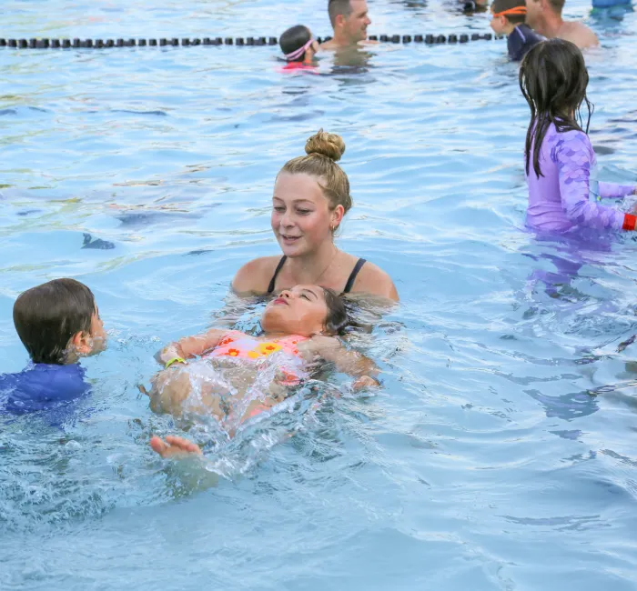 Woman instructor helps a child float on their back in a crowded swimming pool.