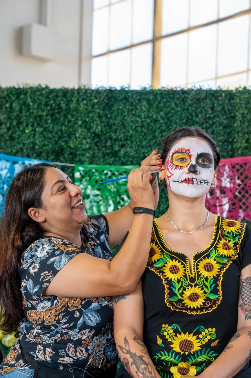 Artist applying vibrant sugar skull makeup to woman's face; festive Day of the Dead celebration.