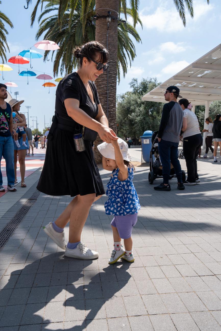 Woman helps a toddler walk on a sunny outdoor path with palm trees and colorful umbrellas.