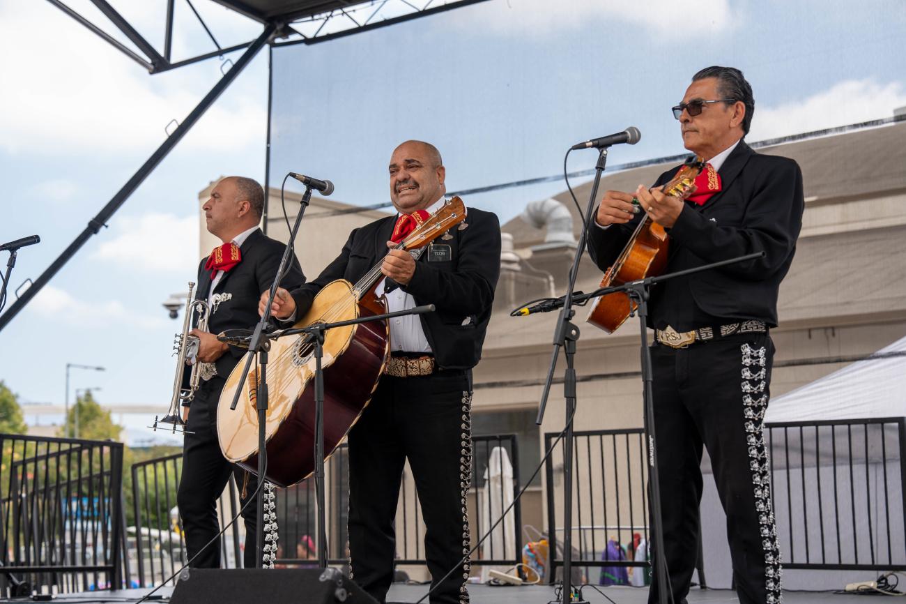 Three mariachi musicians playing guitars on an outdoor stage.