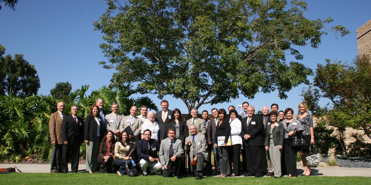 Large group of people in business attire standing on a sunny lawn with trees.