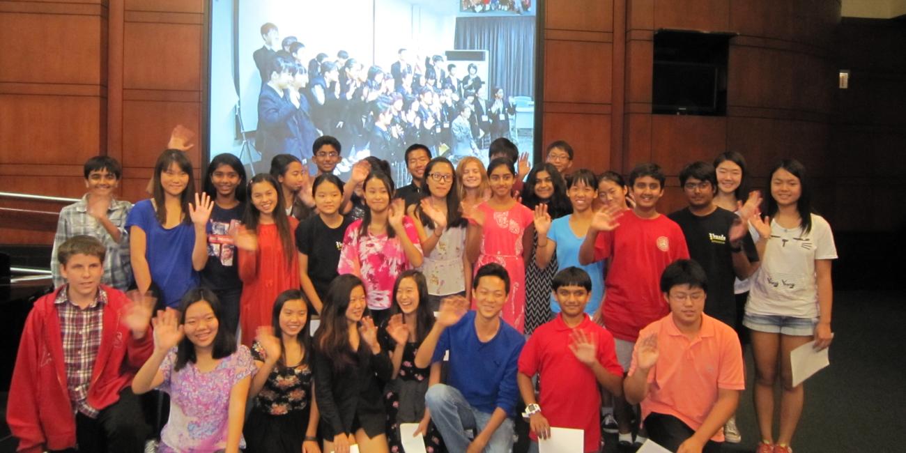 Diverse group of smiling young students waving, with a projected group photo behind them.