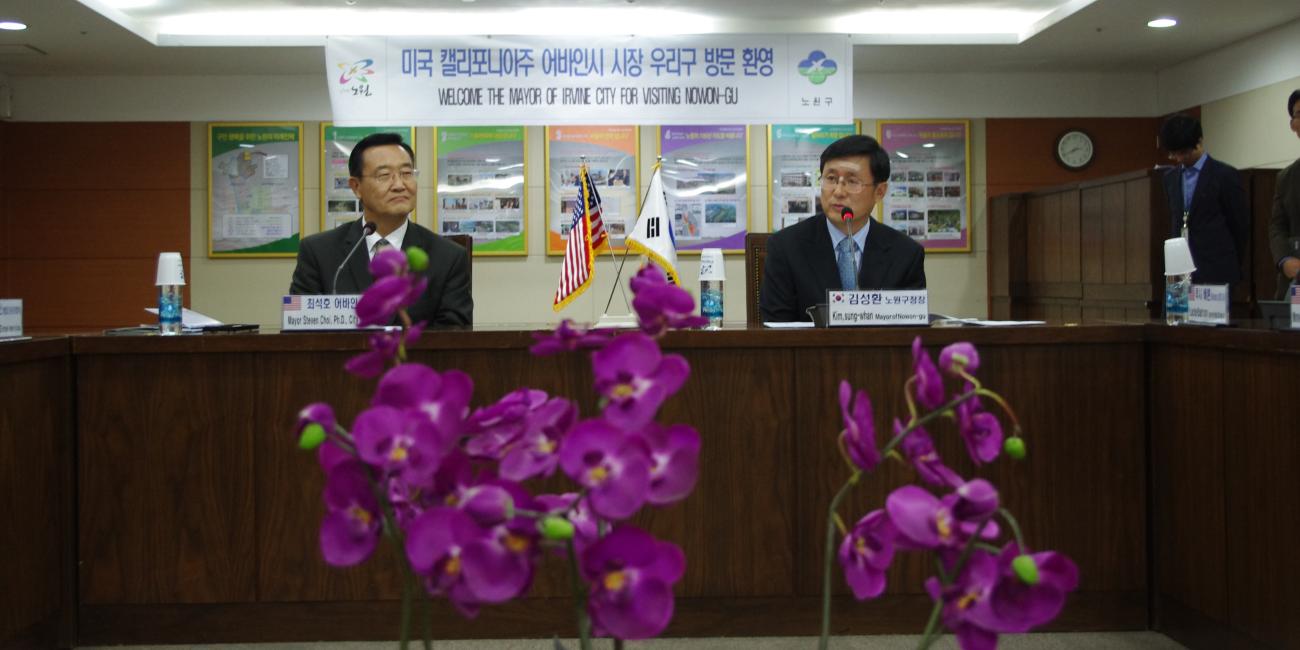 Two men in suits at a conference table, one speaking, with purple orchids and a banner. 