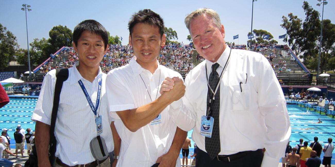 Three smiling men shake hands at an outdoor swim meet.
