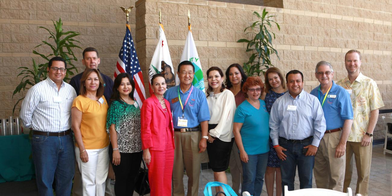 Smiling group of 15 adults, men and women, standing indoors near US and California flags.