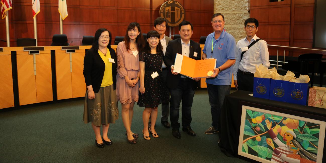 Group of adults in a formal room. Smiling man holds orange folder. Abstract painting on table.