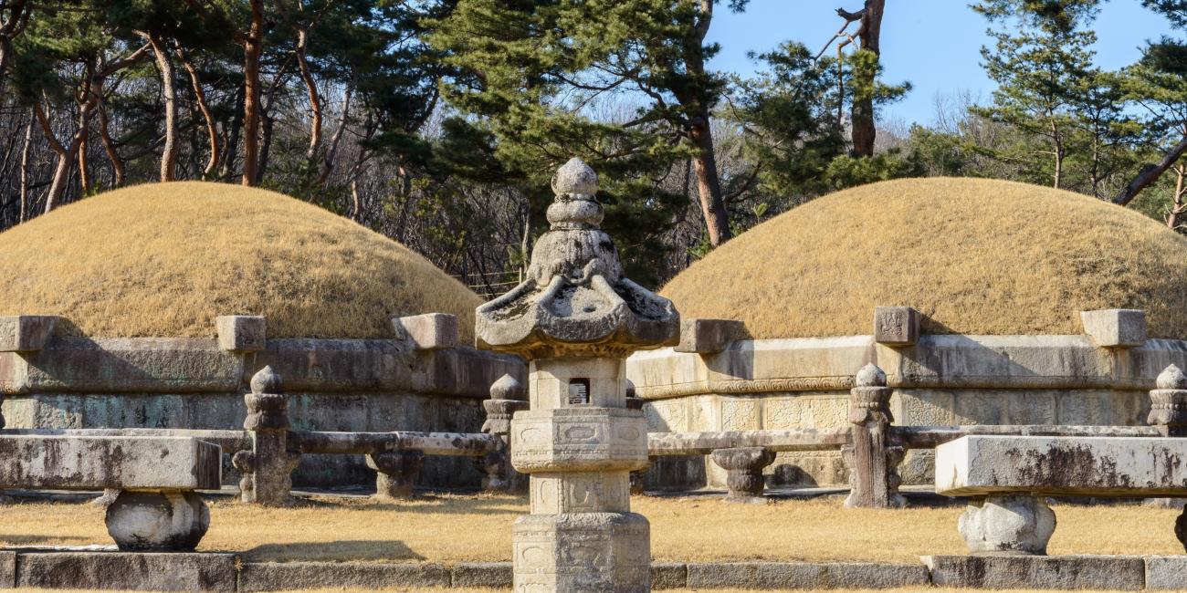 Two ancient grassy burial mounds with ornate stone monuments and pine trees.
