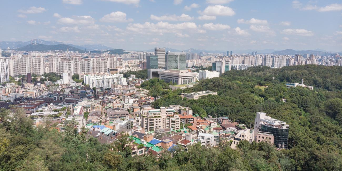 Panoramic city view with lush green hills, residential buildings, and a distant skyline under a blue sky.
