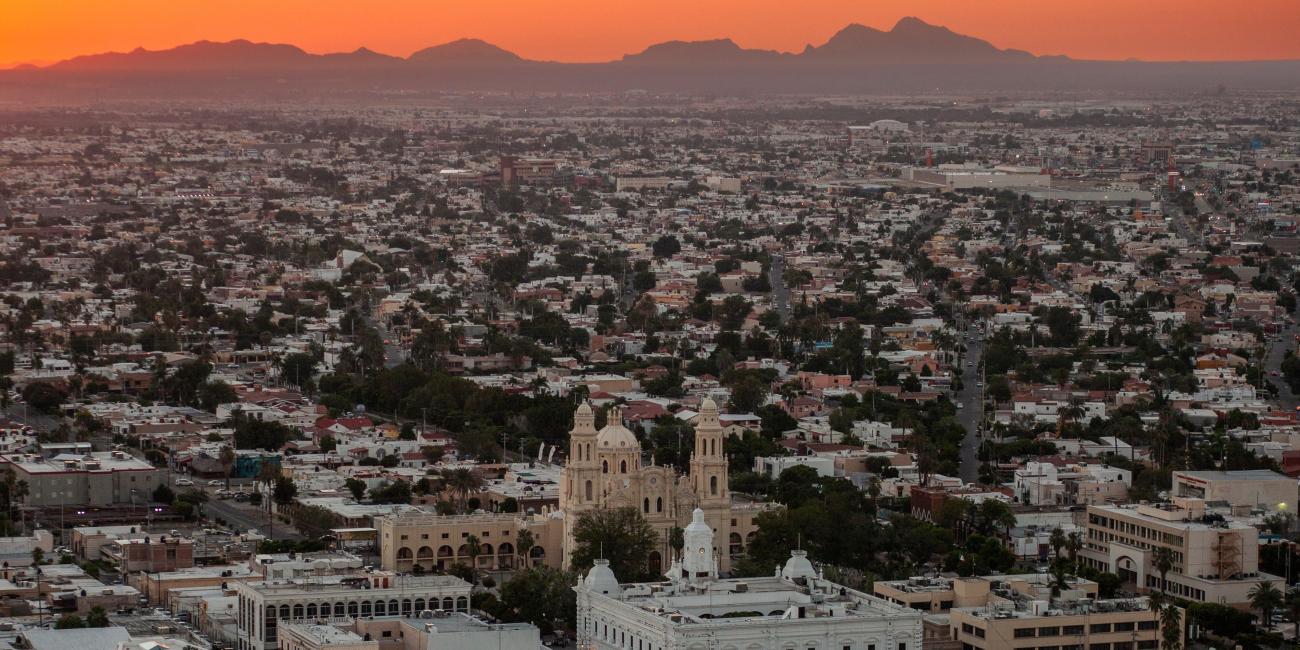 Photograph: Aerial city view at sunset, featuring a large church and mountains under an orange sky.