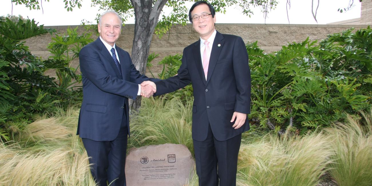 Photograph of two men in suits shaking hands outdoors by a commemorative stone.