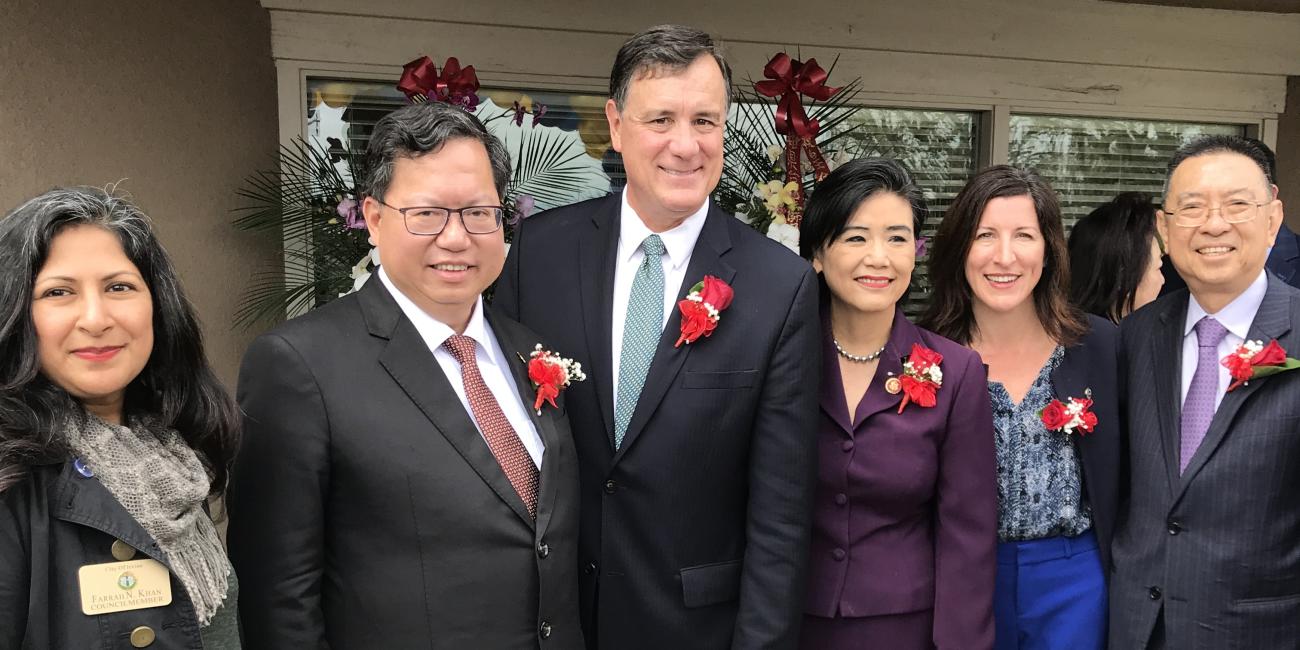 Six smiling people in formal attire with red rose boutonnieres pose for a photograph outdoors.