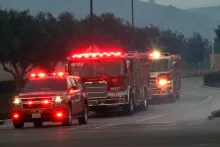 A line of fire trucks with flashing red lights on a road at dusk.