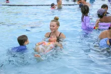 Young woman smiles, supporting a child floating on their back in a swimming pool.
