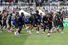 Soccer players in dark uniforms run across a green field with a stadium crowd behind them.
