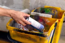 Hand places a spray paint can into a yellow, paint-splattered bucket.