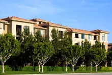 Tan apartment building with red tile roof, lush green trees, and blue sky.