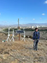 Person stands near scientific instruments and solar panel on a dry hill overlooking a city.