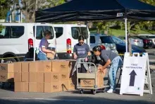 Three people move stacks of cardboard boxes under a black tent in a parking lot.