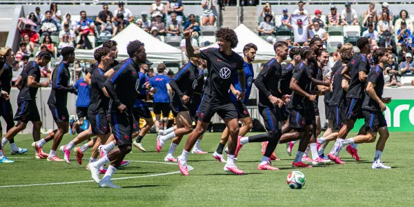 Soccer players in dark uniforms run across a green field with a stadium crowd behind them.