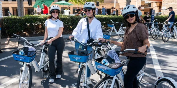 Three women wearing helmets and sunglasses pose with white and blue rental bikes.