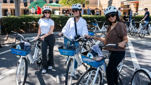 Three women wearing helmets and sunglasses pose with white and blue rental bikes.