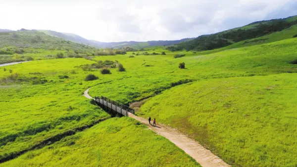 A dirt path winds through bright green hills to a small wooden bridge with two people walking.
