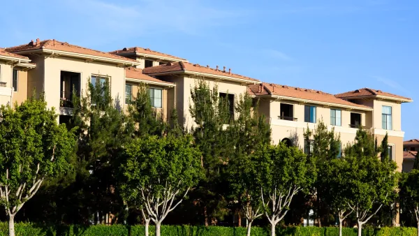 Tan apartment building with red tile roof, lush green trees, and blue sky.