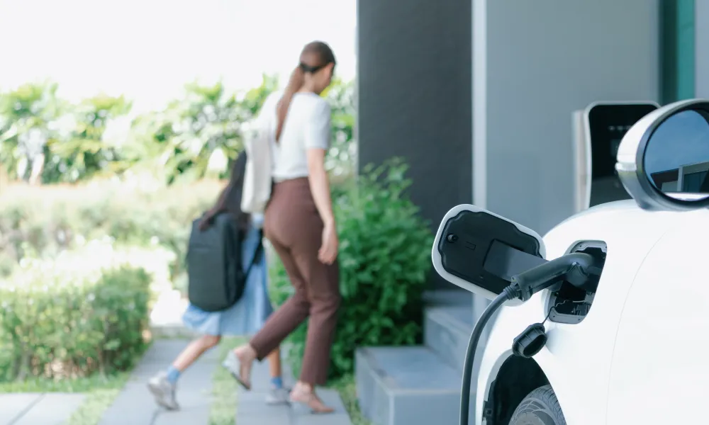 White electric car charging outside a house; woman and child walk inside.