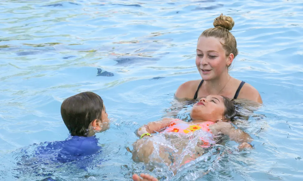 Young woman smiles, supporting a child floating on their back in a swimming pool.