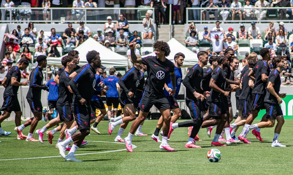 Soccer players in dark uniforms run across a green field with a stadium crowd behind them.