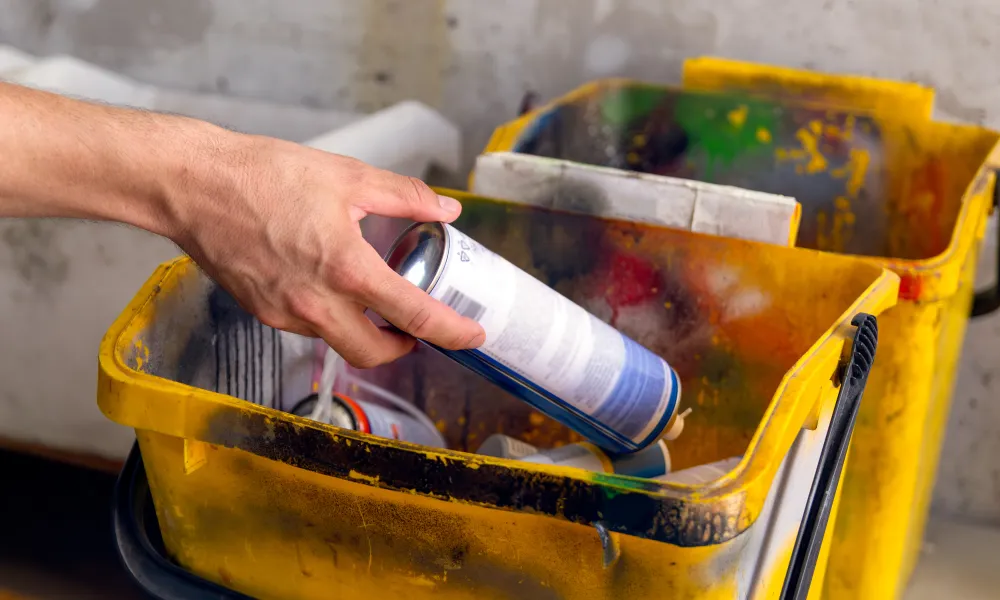 Hand places a spray paint can into a yellow, paint-splattered bucket.