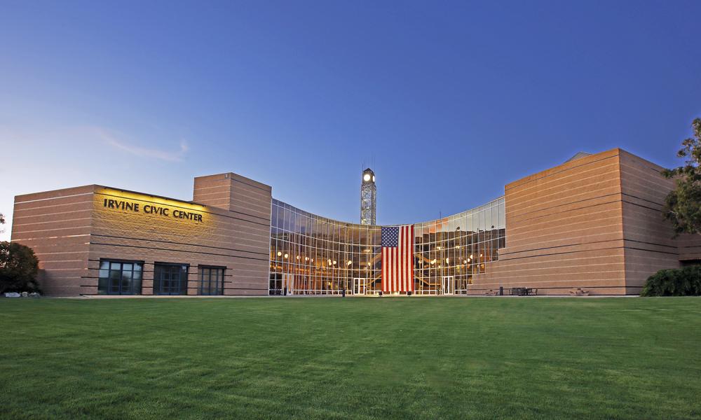Modern Irvine Civic Center with glass facade, clock tower, and American flag over green lawn.