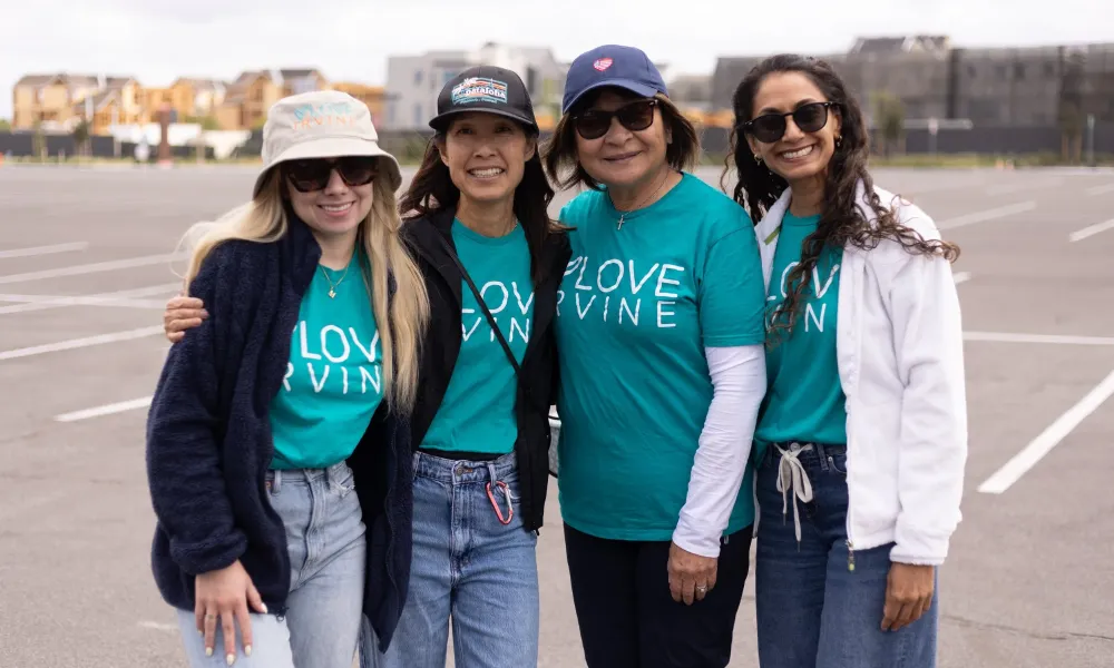 Four smiling women in teal "LOVE IRVINE" shirts stand in a parking lot.