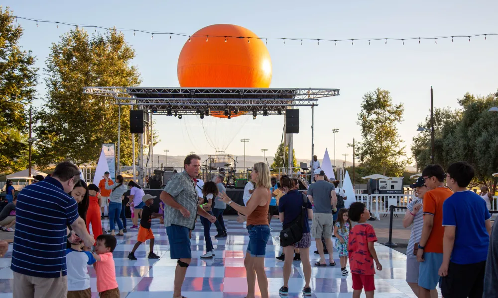 Outdoor event with people dancing on a checkered floor, large orange sphere behind stage.
