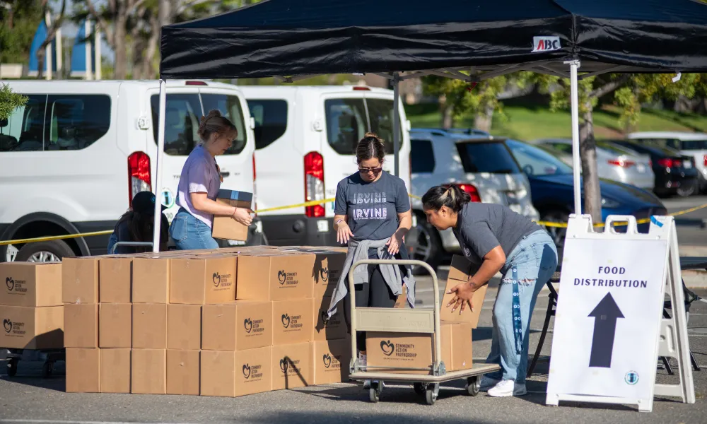 Three people move stacks of cardboard boxes under a black tent in a parking lot.