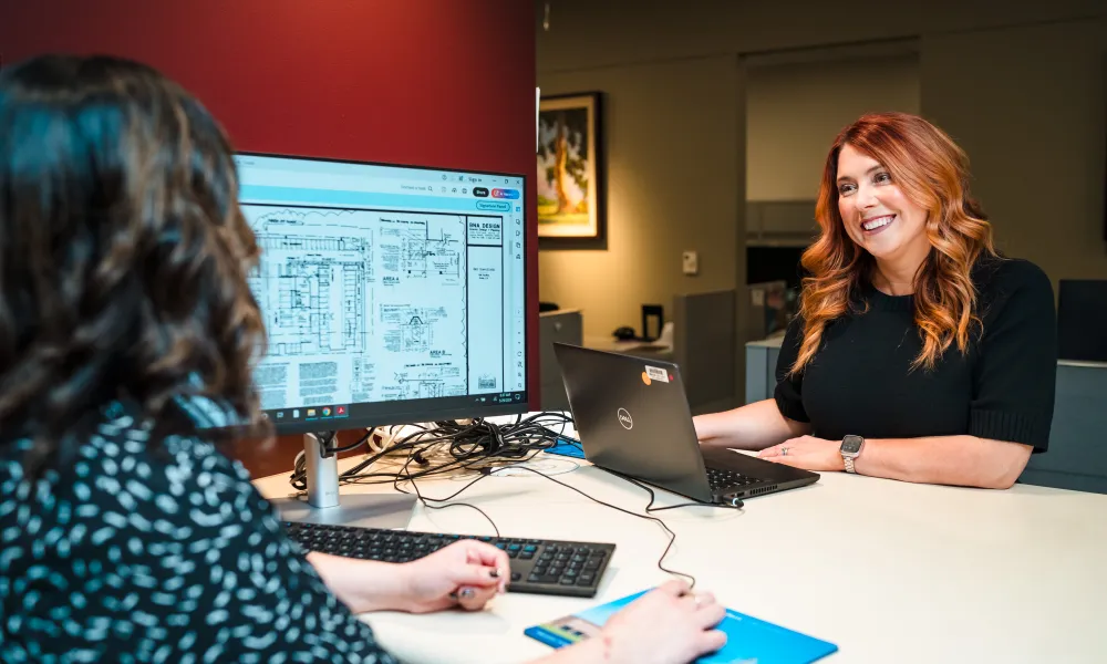 Two women are talking at a desk with a computer.