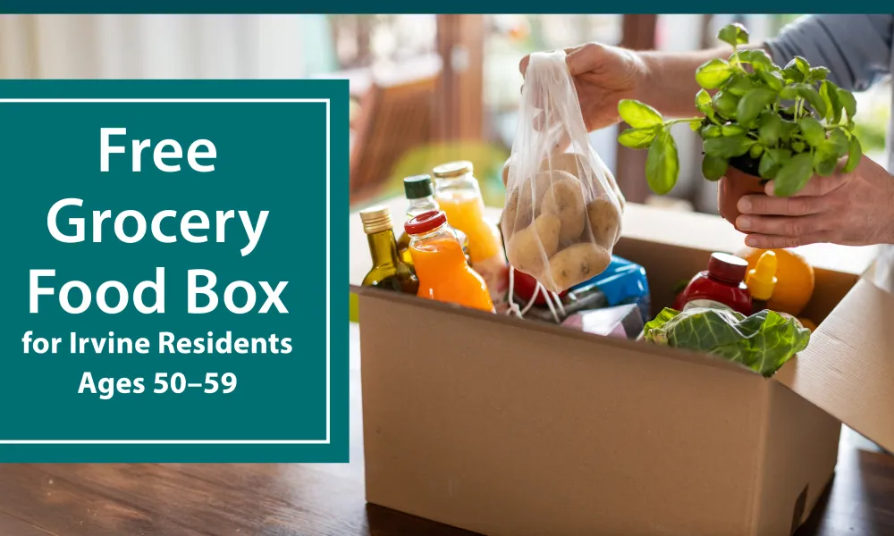 Person's hands filling a cardboard box with fresh groceries on a wooden table.