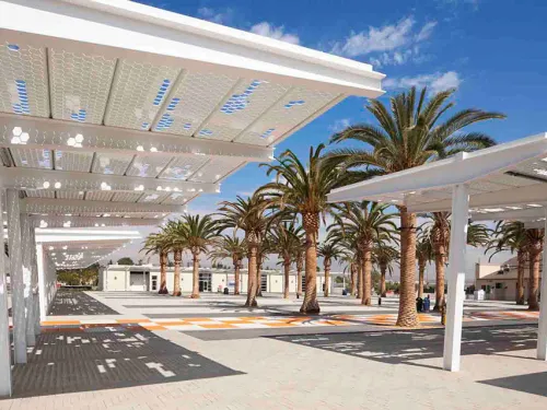 Photograph: Modern white shade structures with patterned roofs, surrounded by palm trees.