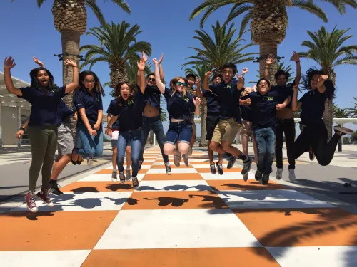 Happy diverse young people jumping on a large orange and white checkerboard path under palm trees.