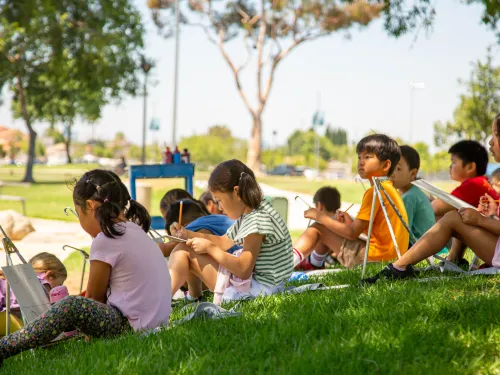 Diverse children drawing on easels in a bright, grassy park.