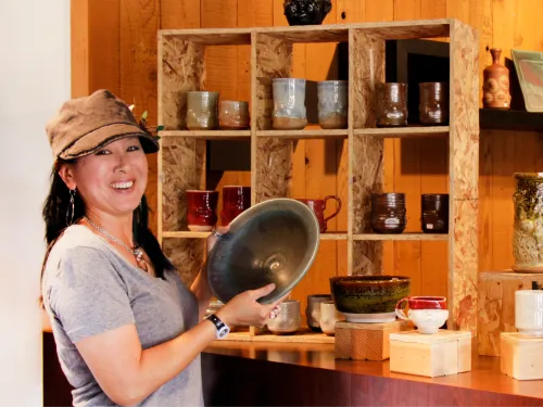 Smiling woman in a hat holds a dark bowl in a pottery shop filled with ceramics.
