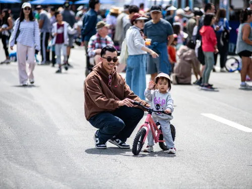 Smiling man helps a toddler on a pink balance bike on a sunny street with many people.