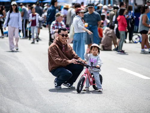 Smiling man helps a toddler on a pink balance bike on a sunny street with many people.